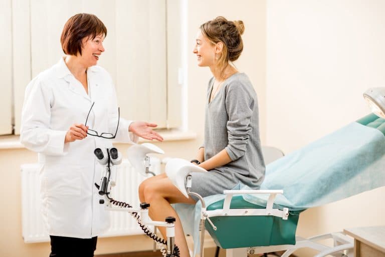 Mulher em consulta ginecológica conversando com médica ao lado da cadeira de exame, representando o diagnóstico e tratamento da endometriose.