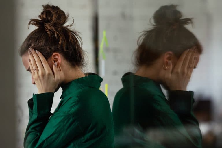 Mulher com as mãos no rosto e expressão de exaustão emocional, refletida em uma superfície espelhada, representando os impactos do burnout na saúde mental, no estresse crônico e no esgotamento relacionado ao trabalho.