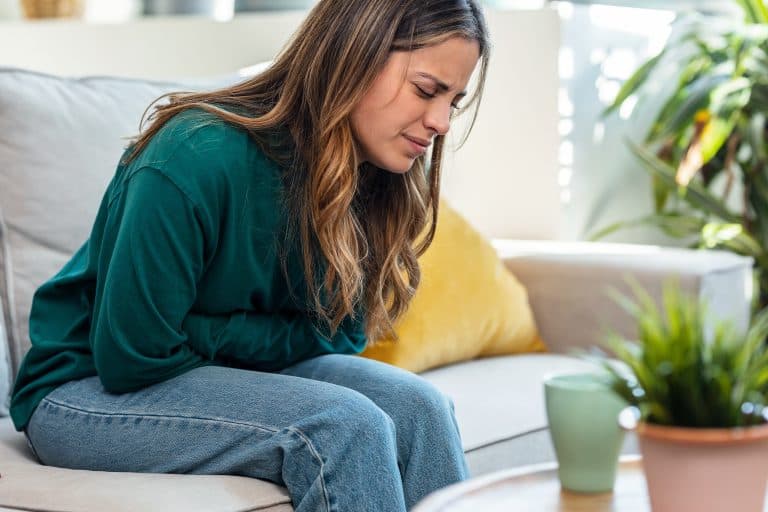 Mulher sentada no sofá com as mãos sobre o abdômen e expressão de desconforto, ilustrando sintomas da doença celíaca, como dor abdominal, inchaço e alterações digestivas que exigem avaliação médica especializada.
