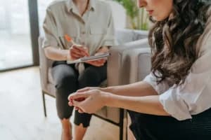 Mulher em atendimento psicológico, com expressão preocupada, enquanto profissional faz anotações, representando a importância da saúde mental durante a busca por emprego.