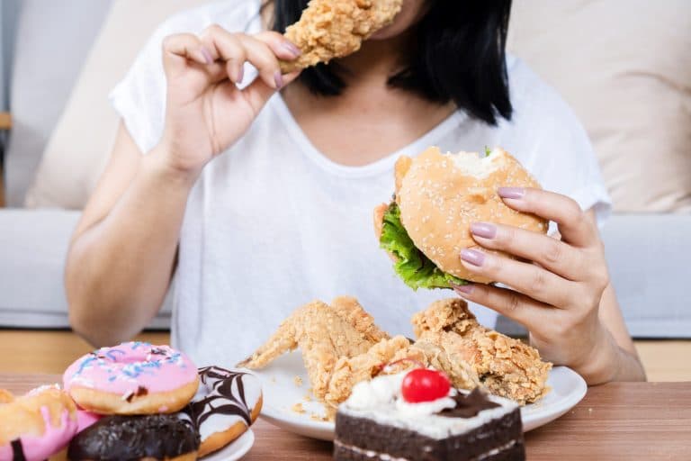 Pessoa consumindo hambúrguer, frango frito e doces, ilustrando o comer emocional, quando a alimentação é usada para aliviar emoções em vez de saciar a fome.
