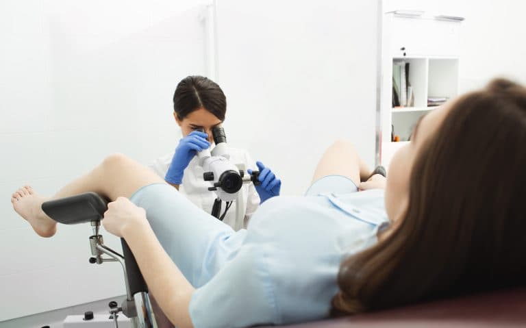 Paciente realiza exame ginecológico em consultório, com profissional de saúde utilizando equipamento para avaliação do colo do útero.